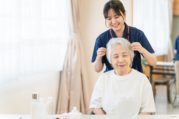 Caregiver helps an elderly woman style her hair at a vanity, both smiling, with toiletries on the counter.