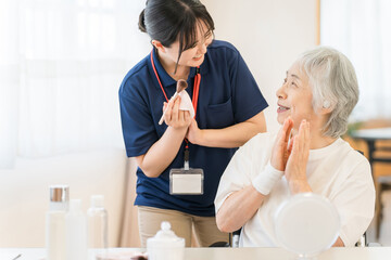Caregiver in blue scrubs with a name badge chats with an elderly woman at a table, both smiling and clapping.