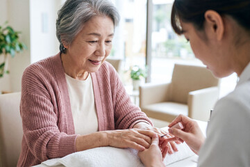Elderly woman with a warm smile receives gentle hand care from a caregiver in a bright living room.