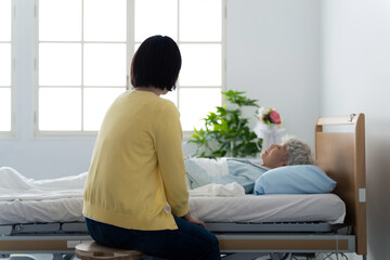 Caregiver seated on a stool beside a hospital bed, talking to an elderly patient in a sunlit room.