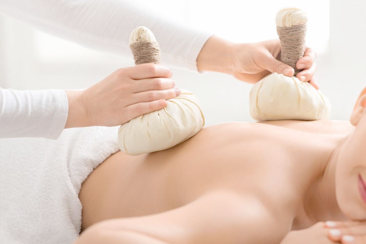 Two hands press warm herbal compresses on a person's back during a spa massage session.