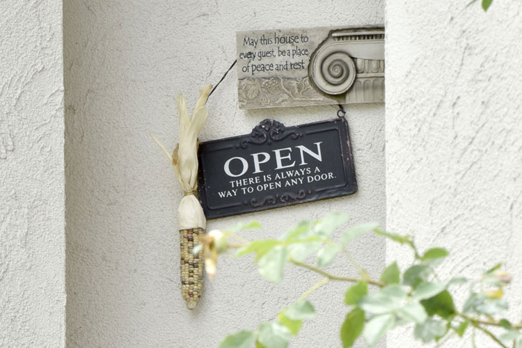 Open sign with the text 'OPEN THERE IS ALWAYS A WAY TO OPEN ANY DOOR' hanging on a textured wall beside a corn decor and a small peace plaque.