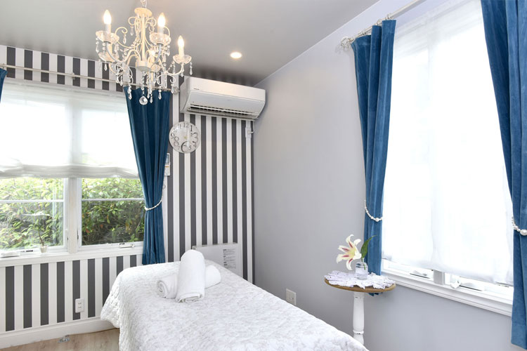 Spa-style treatment room with a white massage bed, folded towels, blue curtains, and a striped accent wall under a crystal chandelier