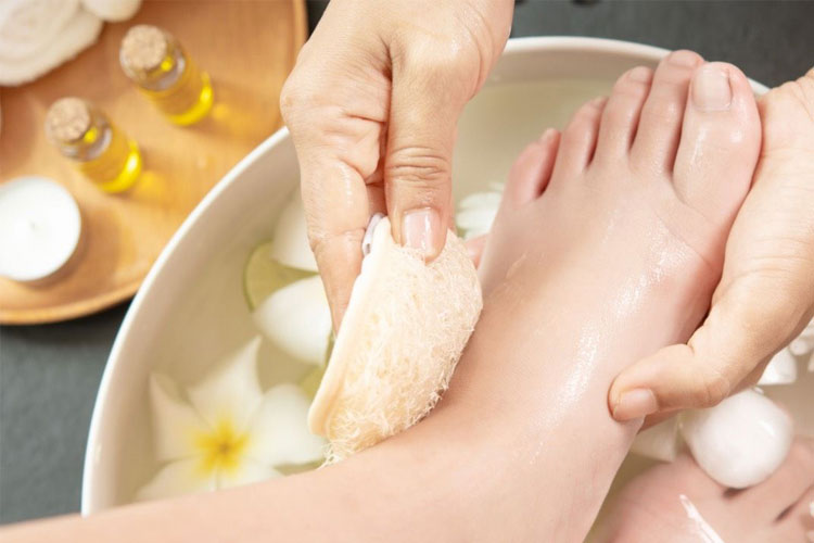 Foot being exfoliated with a loofah during a foot spa pedicure in a bowl of water with petals and oils nearby.