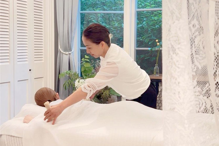 Massage therapist giving a back massage to a client lying on a table in a bright, airy room with plants nearby.