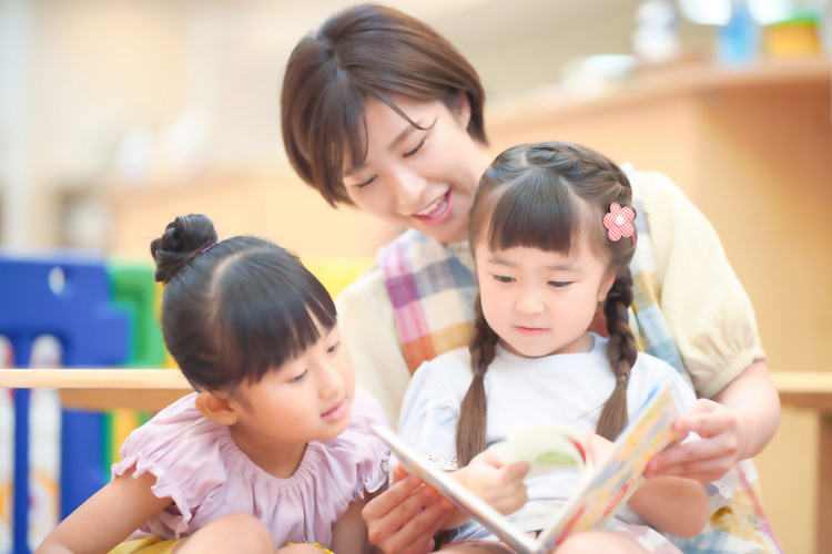 Caregiver reading a storybook to two young girls in a bright classroom, all focused on the book