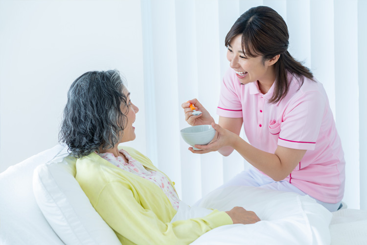 Caregiver in a pink uniform feeds an elderly woman sitting up in bed, using a bowl and spoon.