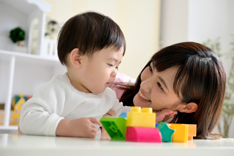 Mother and baby smiling at each other while playing with colorful building blocks on a white table.