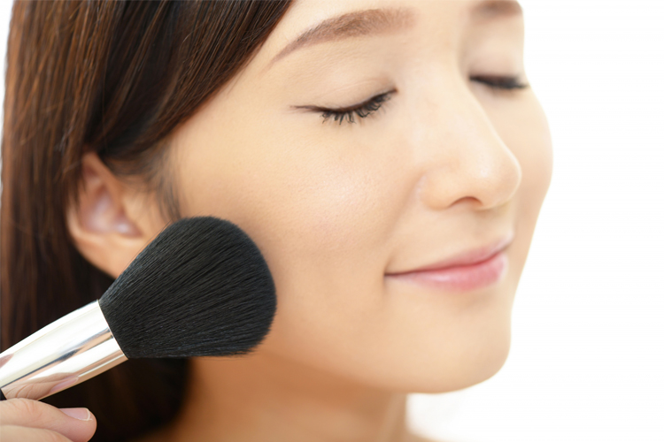 Woman smiling as she applies makeup with a large brush on her cheek.